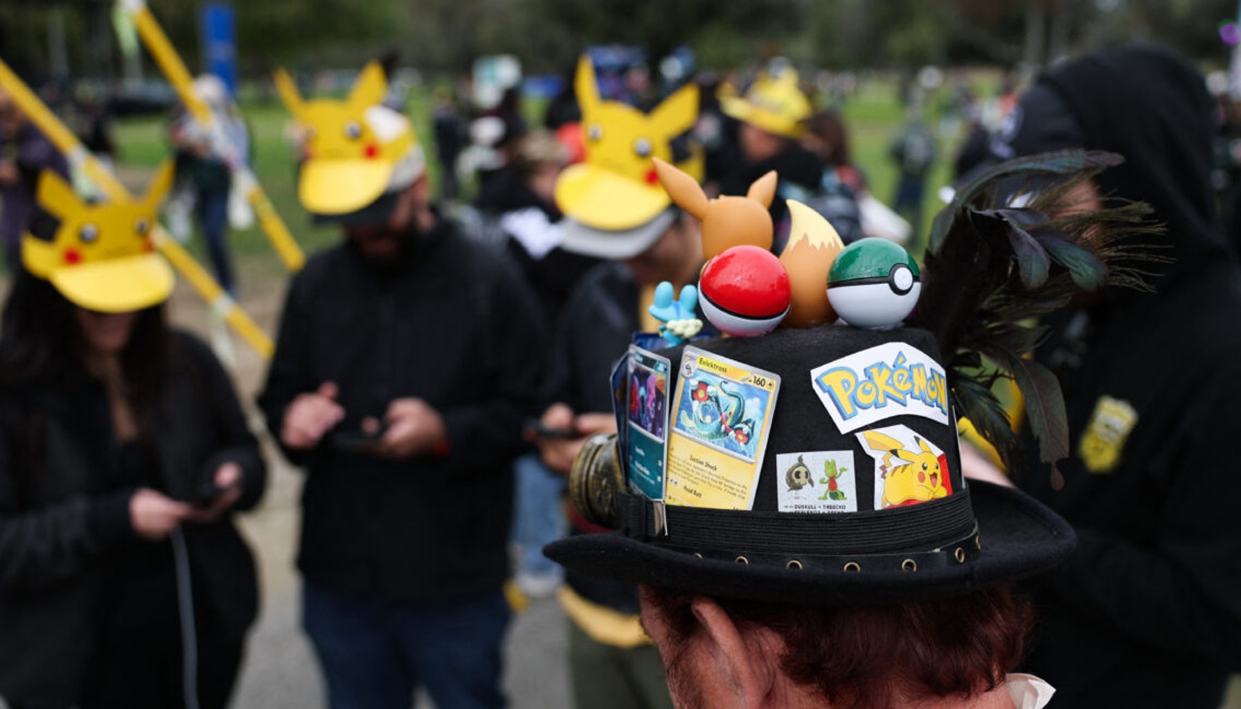 A player wearing a hat decorated with Pokemon characters and trading cards plays Pokemon GO on a smartphone during the in-person Pokemon GO Tour: Kalos Los Angeles 2026 event at the Rose Bowl Stadium in Pasadena, California, on February 20, 2026.