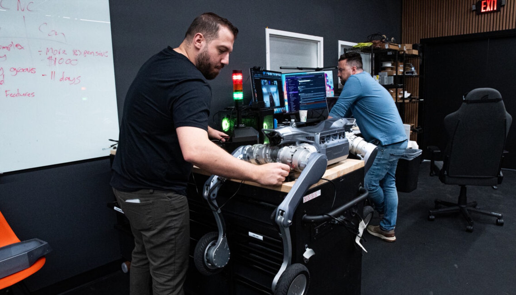 A person works on a quadruped robotic dog that is lying on a counter, while a second person stands in front of a computer with multiple display screens inside the office of Robo Inc.