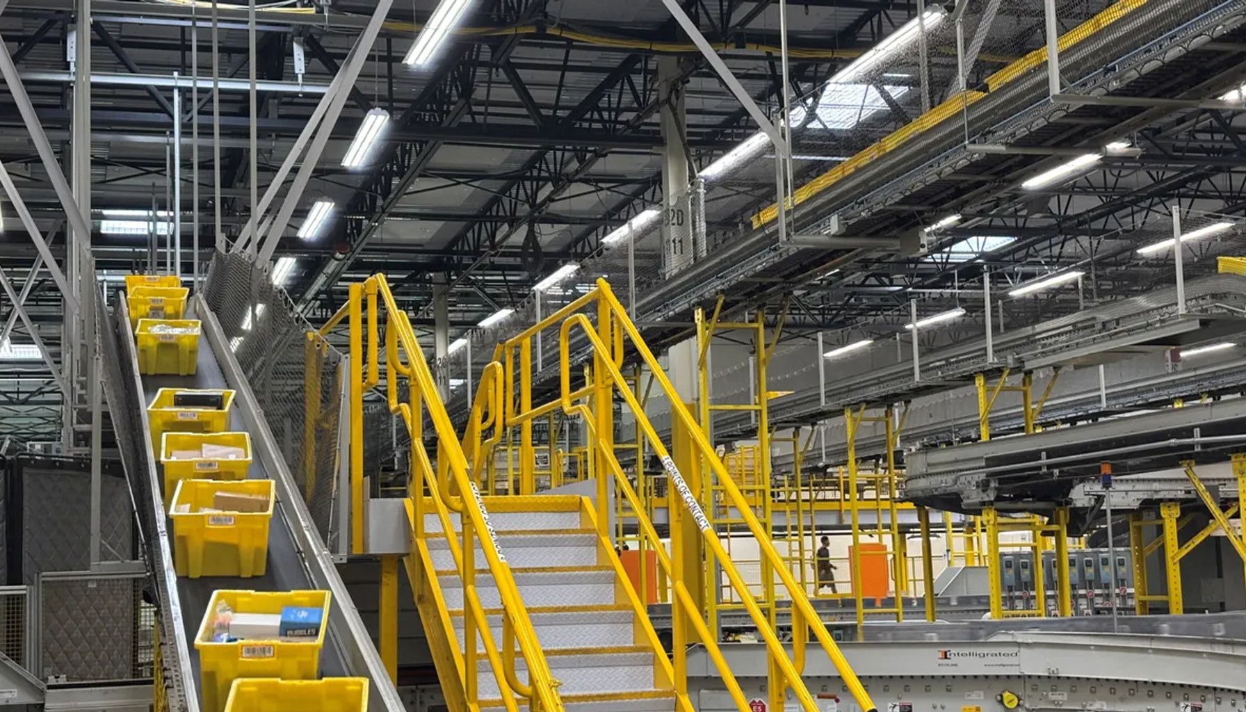 Yellow bins move on a conveyor belt located next to a set of stairs with yellow railings within a warehouse.